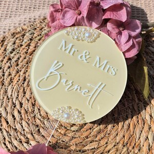 May include: A round, pale yellow wedding cake topper with the text "Mr & Mrs Barnett" in white script. The topper is decorated with pearl accents and is surrounded by pink flowers.