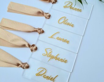 May include: Five clear acrylic place cards with gold lettering. The names Deanna, Olivia, Lauren, Stephanie, and Daniel are written on the cards. Each card has a beige satin ribbon tied to it.