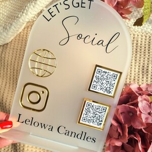 May include: A white frosted acrylic sign with the text "Let's Get Social" in black lettering. The sign features a gold globe icon, a gold Instagram icon, and two gold framed QR codes. The text "Lelowa Candles" is printed in black at the bottom of the sign.