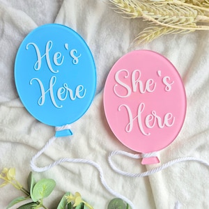 May include: Two round, balloon-shaped signs with white strings attached. The blue sign says "He's Here" and the pink sign says "She's Here".