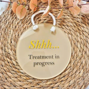 May include: A round, tan sign with the text "Shhh... Treatment in progress" in yellow and black. The sign has a white handle and is set against a woven background with peach-colored leaves.