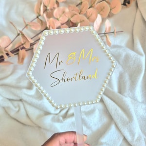 May include: A hexagonal wedding cake topper with a pearl border. The topper has gold lettering that reads "Mr & Mrs Shortland". The topper is held by a clear plastic stick. The background includes pink leaves and a white cloth.