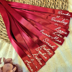 May include: A collection of long, deep red satin ribbons, each featuring a name printed in white cursive script. The names visible on the ribbons include Brooke, Beverley, Lizzie, Lisa, Laurie-Mae, Sarah, Lesley, Sophie, and Anne.