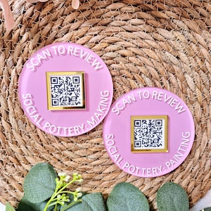 May include: Two pink round coasters with white text that reads "Scan to Review Social Pottery Making" and "Scan to Review Social Pottery Painting". Each coaster has a gold square with a black and white QR code in the center.