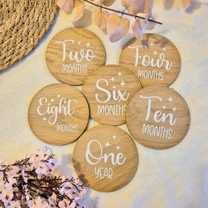May include: A collection of six wooden milestone discs, each with white lettering indicating a baby's age in months or years. The discs are labelled: Two, Four, Six, Eight, Ten Months, and One Year.