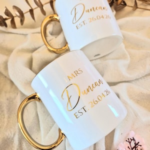 May include: Two white ceramic mugs with gold handles. The mugs have gold lettering that reads "MR Duncan EST. 26.04.26" and "MRS Duncan EST. 26.04.26". The mugs are on a white cloth.