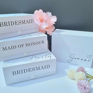 May include: Three white gift boxes with black text. The top box says "BRIDESMAID", the middle box says "MAID OF HONOUR", and the bottom box says "BRIDESMAID" with the name "Carly" written above it. A fourth white box says "Joanne" with a heart symbol. The boxes are on a gray surface with pink and white flowers.