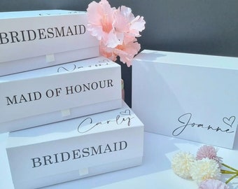 May include: Three white gift boxes with black text. The top box says "BRIDESMAID", the middle box says "MAID OF HONOUR", and the bottom box says "BRIDESMAID" with the name "Carly" written above it. A fourth white box says "Joanne" with a heart symbol. The boxes are on a gray surface with pink and white flowers.