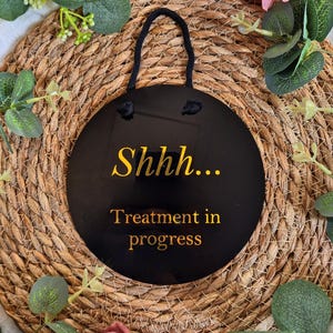 May include: A black, round sign with the text "Shhh... Treatment in progress" in gold lettering. The sign has a black rope for hanging. The sign is surrounded by greenery and pink flowers.