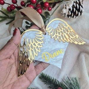 May include: A heart-shaped Christmas decoration with gold angel wings and the name "Diane" in yellow script. The ornament has a white and grey marbled pattern and is suspended by a beige ribbon. Festive greenery and red berries are in the background.