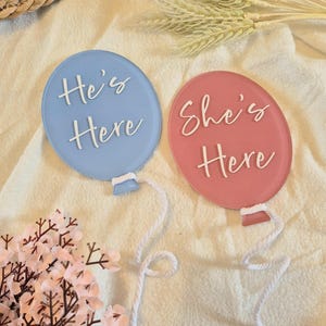 May include: Two balloon-shaped signs, one baby blue and one pink, with the text "He's Here" and "She's Here" in white script. Each balloon has a white string attached. The signs are on a cream-coloured surface, with floral and decorative elements.