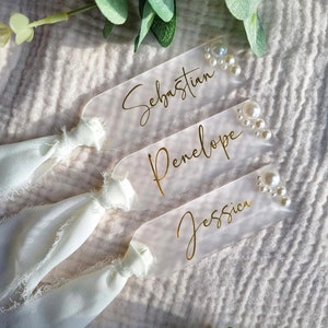 May include: Three clear acrylic place cards with gold calligraphy names "Sebastian", "Penelope", and "Jessica". Each card has a cluster of white pearl embellishments.
