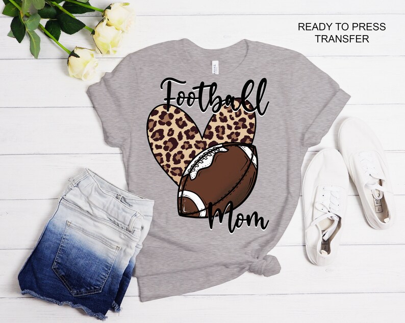 Pu&ograve; includere: Maglietta grigia con un cuore a stampa leopardata e un design di pallone da calcio. Il testo "Football Mom" &egrave; stampato sulla maglietta.
