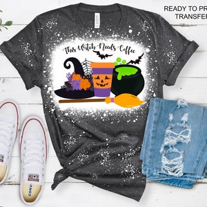 May include: A dark gray t-shirt with a white splatter design. The shirt has a graphic design of a witch's hat, a coffee cup with a jack-o-lantern face, a cauldron, and a broom. The text "This Witch Needs Coffee" is printed on the shirt.