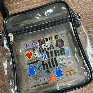 May include: Clear plastic crossbody bag with black trim and a zipper closure. The bag is decorated with various stickers, including the words "One Tree Hill", "Scott", "3", "23", and "Ravens".