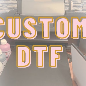 May include: A close-up of a printer with the words "CUSTOM DTF" in pink and gold letters. There are several bottles of digital ink in the foreground.