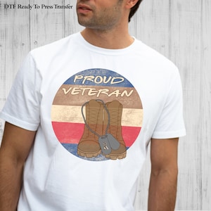 May include: A white t-shirt with a circular design featuring a pair of brown boots with dog tags, a red, white, and blue striped background, and the text "PROUD VETERAN".