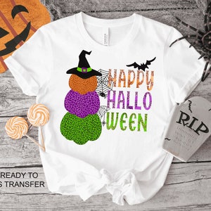 May include: White t-shirt with a graphic of a stack of pumpkins with a witch's hat on top. The pumpkins are green, purple, and orange with black spots. The text "Happy Hallo Ween" is above the pumpkins.