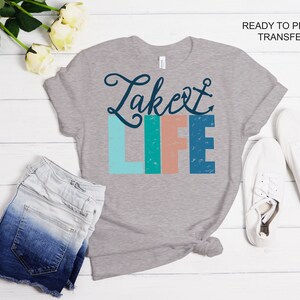 May include: Ready to press iron-on transfer design featuring the text "Lake Life" in a colorful, distressed font with an anchor symbol. The design is on a gray t-shirt with a pair of white sneakers and denim shorts in the background.
