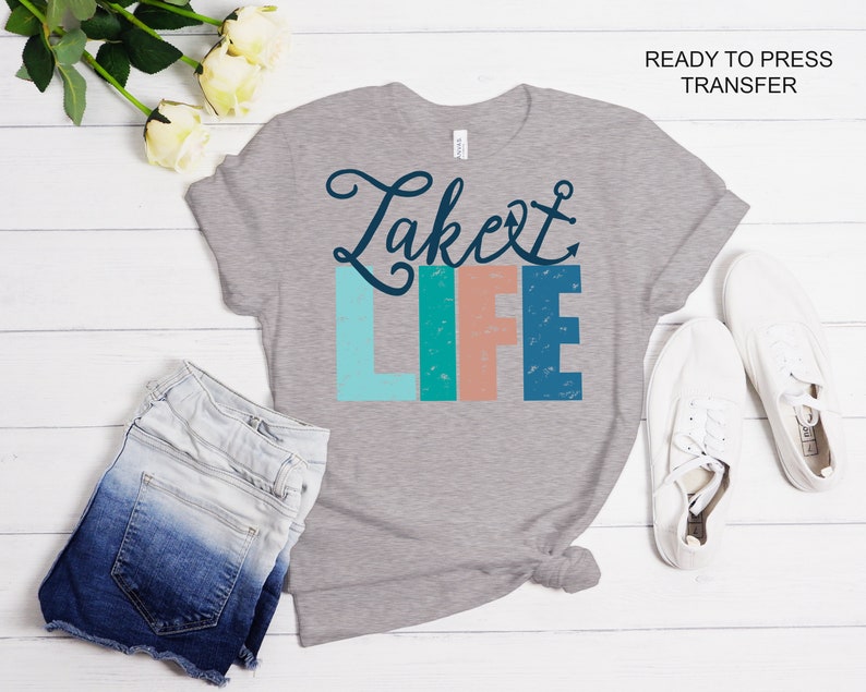May include: Ready to press iron-on transfer design featuring the text "Lake Life" in a colorful, distressed font with an anchor symbol. The design is on a gray t-shirt with a pair of white sneakers and denim shorts in the background.