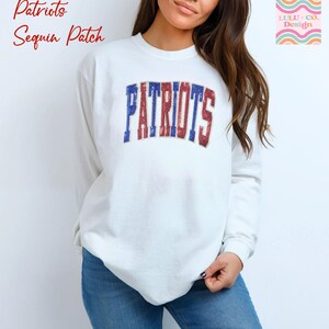 May include: A white long-sleeve sweatshirt with the word "PATRIOTS" in blue and red sequin letters across the chest. The sweatshirt also has the text "Patriots Sequin Patch" in red script in the upper left corner.