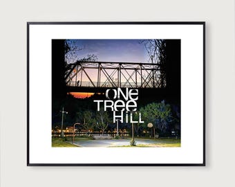 May include: Framed print featuring a bridge silhouette against a sunset sky, with the text "ONE TREE HILL" overlaid. The lower portion shows a basketball court and trees, lit by streetlights, creating a scenic landscape.