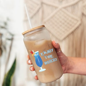 May include: A glass iced coffee drink in a clear glass with a bamboo lid and a straw. The glass has a blue and white sticker with a cocktail glass and the text "Enjoy the Buzz!"