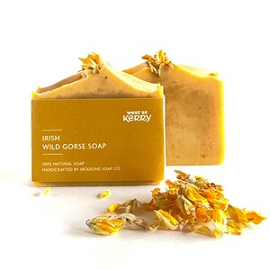 Könnte beinhalten: Ein rechteckiges Stück gelbe Irish Wild Gorse Seife, eingewickelt in ein senffarbenes Papieretikett mit weißem Text: West of Kerry, Irish Wild Gorse Soap, 100% Natural Soap, Handcrafted by Mckeone Soap Co. Daneben liegen getrocknete gelbe Blütenblätter.