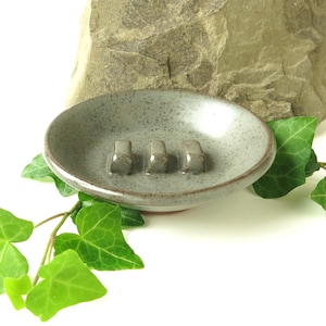 May include: A round, speckled gray ceramic soap dish with a smooth, glazed finish. The interior features three raised, rounded ridges designed to elevate a bar of soap, while the outer rim is defined by a thin, dark brown edge.