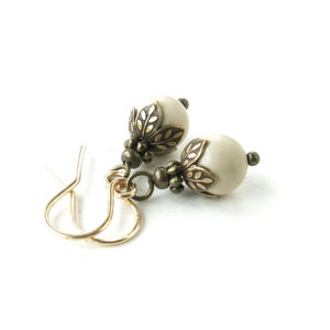May include: A pair of cream-colored beaded earrings with gold-colored ear wires. Each earring features a round, cream-colored bead with antique gold-colored leaf-shaped accents and smaller beads. The earrings are set against a white background.
