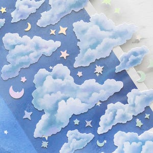 Puede incluir: Una hoja de pegatinas titulada "COZY CLOUDS" con nubes azules de acuarela, estrellas y lunas crecientes. Las pegatinas tienen un brillo holográfico. El fondo es un degradado de tonos azules.