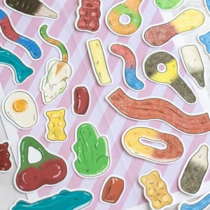 Op de afbeelding: Assortiment van kleurrijke gummy snoepjes in verschillende vormen, waaronder een kikker, een kers, een donut en een werveling. De snoepjes zijn gerangschikt op een roze en witte gestreepte achtergrond.