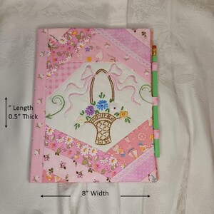 May include: A pink quilted journal with an embroidered floral basket design. The journal is 8 inches wide and 0.5 inches thick. A green pencil is attached to the side. The cover features various pink floral patterns.