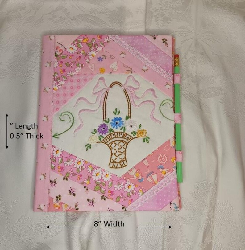May include: A pink quilted journal with an embroidered floral basket design. The journal is 8 inches wide and 0.5 inches thick. A green pencil is attached to the side. The cover features various pink floral patterns.