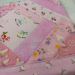 May include: A patchwork textile in shades of pink, featuring various patterns including polka dots, floral prints, and illustrations of kittens, birds, and sailboats. The edges are finished with a delicate lace trim, creating a charming, vintage aesthetic.