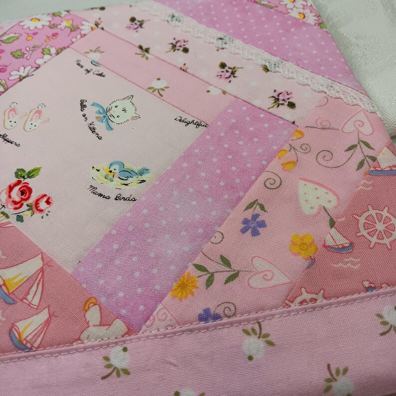 May include: A patchwork textile in shades of pink, featuring various patterns including polka dots, floral prints, and illustrations of kittens, birds, and sailboats. The edges are finished with a delicate lace trim, creating a charming, vintage aesthetic.