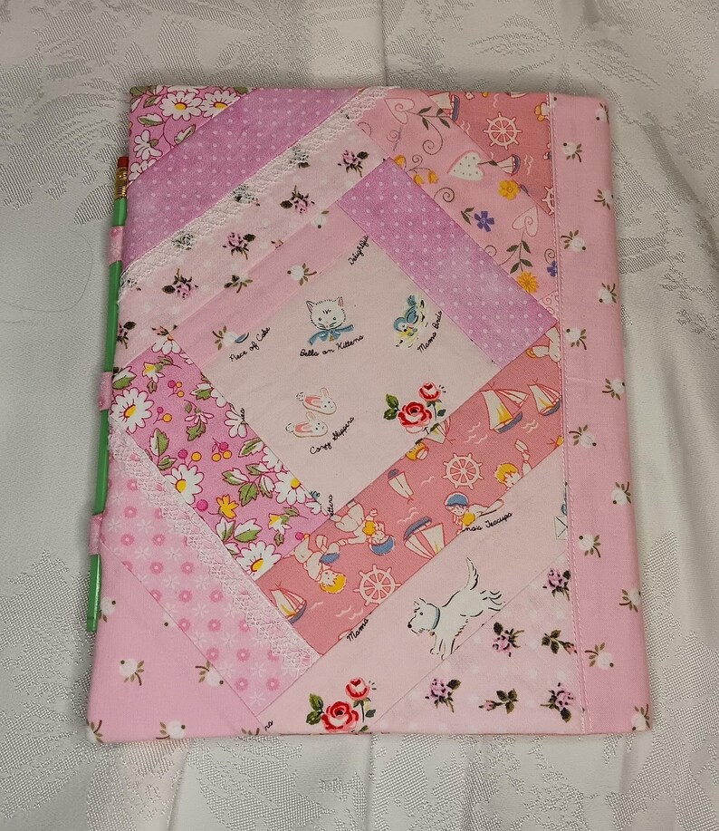 May include: A pink and white quilted notebook cover with a variety of patterns. The cover features floral, polka dot, and nautical-themed fabric patches. The design includes images of kittens, sailboats, and a white dog.