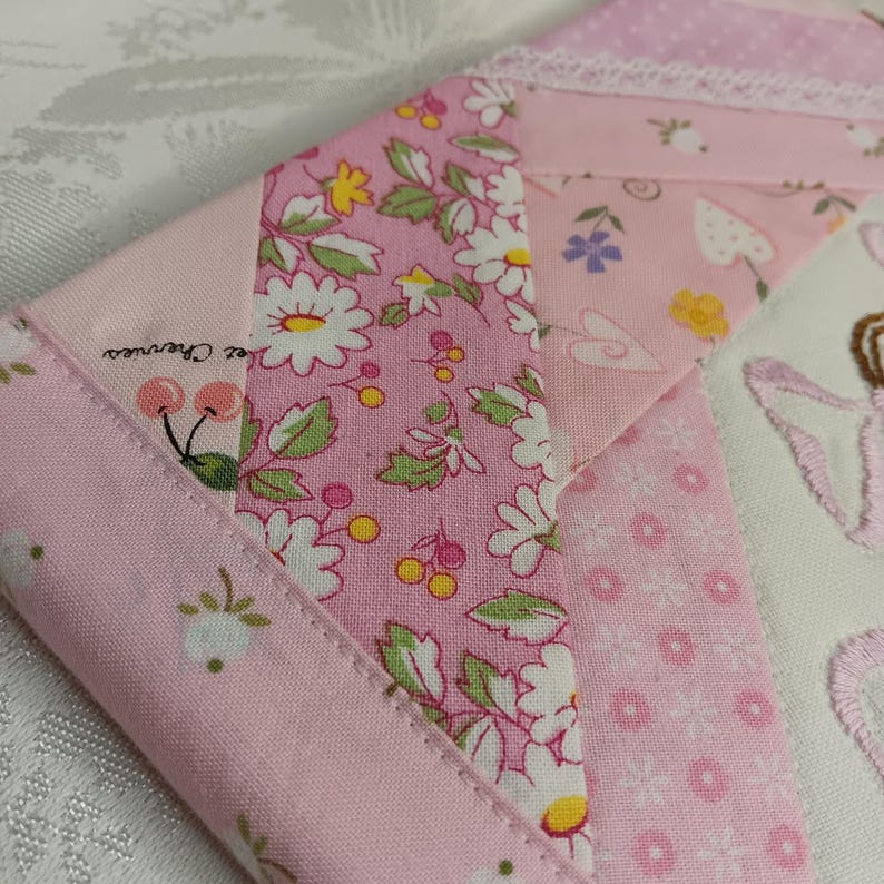 May include: Close-up of a patchwork textile with various pink floral patterns and a white lace trim. The fabric features daisies, cherries, and other small floral designs. The text "Chez Cherries" is visible.