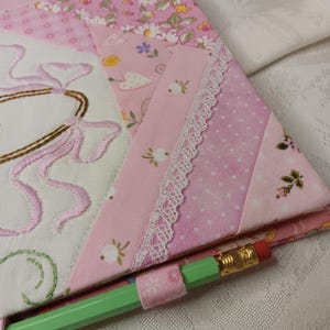 May include: A close-up of a pink and white quilted fabric with floral and polka dot patterns. The fabric is embellished with embroidered details and white lace trim. A green pencil with a pink fabric holder is in the foreground.