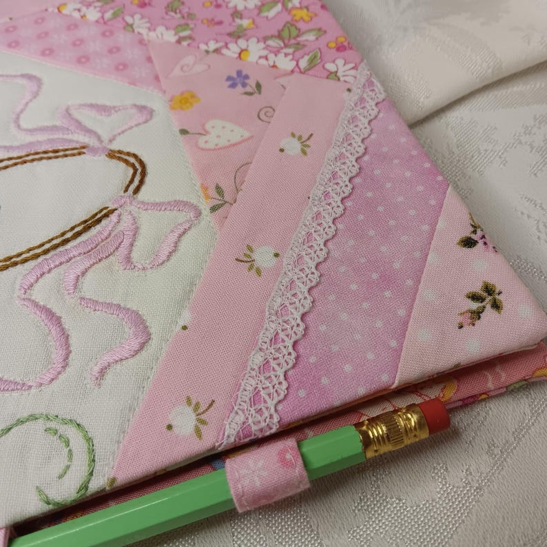 May include: A close-up of a pink and white quilted fabric with floral and polka dot patterns. The fabric is embellished with embroidered details and white lace trim. A green pencil with a pink fabric holder is in the foreground.