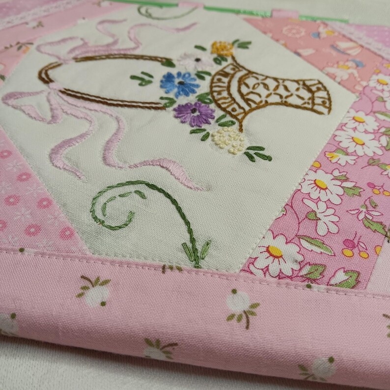 May include: A quilted, floral-themed fabric square with an embroidered design. The central design features a basket of flowers and a pink ribbon. The surrounding fabric is pink with floral patterns and a white background.