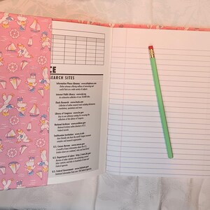 May include: Open notebook with lined pages and a green pencil. The cover is pink with a nautical pattern of sailors, sailboats, and anchors. The left page has a list of search sites.