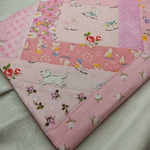 May include: A patchwork quilt square in shades of pink. The fabrics feature floral, nautical, and animal prints, including sailboats, kittens, and a white dog. The edges are finished with a pink floral border, creating a charming, vintage aesthetic.