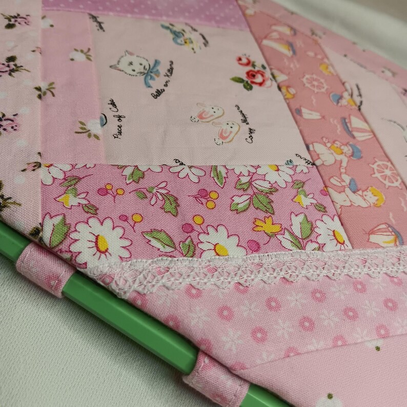 May include: A patchwork fabric item in shades of pink, featuring floral, cat, and nautical patterns. The design includes a white lace trim and a green handle. The fabric is a mix of different patterns and textures.