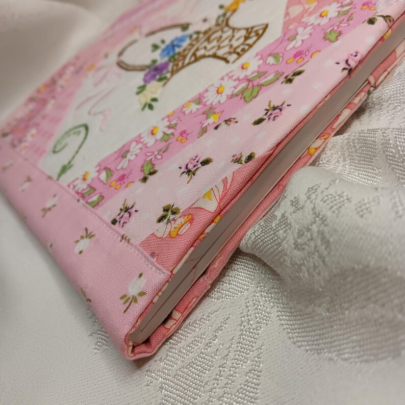 May include: A fabric-covered notebook with a floral design. The cover features a patchwork design with pink and white floral patterns, a basket of flowers, and a pink border. The notebook is closed, showing the edge of the pages.