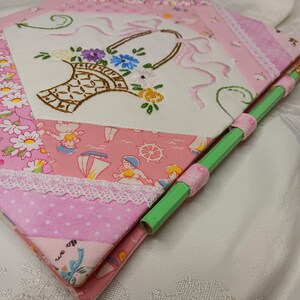 May include: A handmade pink fabric notebook with a floral embroidered design. The notebook features a basket of flowers, lace trim, and a green pencil secured with pink ribbon. The notebook is made of various pink patterned fabrics.