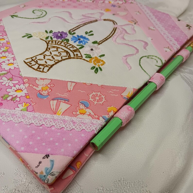 May include: A handmade pink fabric notebook with a floral embroidered design. The notebook features a basket of flowers, lace trim, and a green pencil secured with pink ribbon. The notebook is made of various pink patterned fabrics.