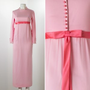 以下が含まれることがあります： 長袖とハイネックの淡いピンク色のヴィンテージドレス。ドレスは、コントラストのあるコーラル色のサテンリボンベルトとウエストの蝶ネクタイが特徴です。小さなボタンが胴体を飾っています。袖口はフリルになっています。