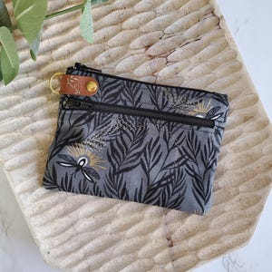 May include: A small, gray zippered pouch with a black and gold leaf and insect pattern. The pouch has a brown leather tag with a gold keyring attached. The pouch is on a carved wooden tray.