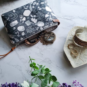 May include: A gray and white fabric pouch with a zipper closure and a brown leather strap. The pouch is decorated with a floral pattern and is filled with jewelry, including a dragonfly pendant, a beaded bracelet, and several rings.